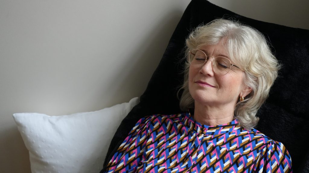 Frau meditiert mit geschlossenen Augen liegend in einem Liegestuhl in einem hellen Raum